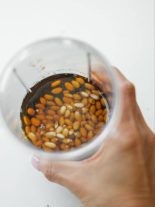 A hand holding a clear cup filled with brown liquid and soaked pine nuts on a white surface.